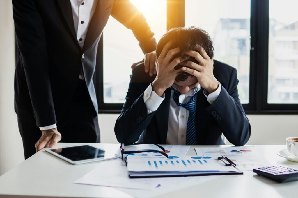 An overwhelmed businessman grasps his head, surrounded by reports in a bright office.